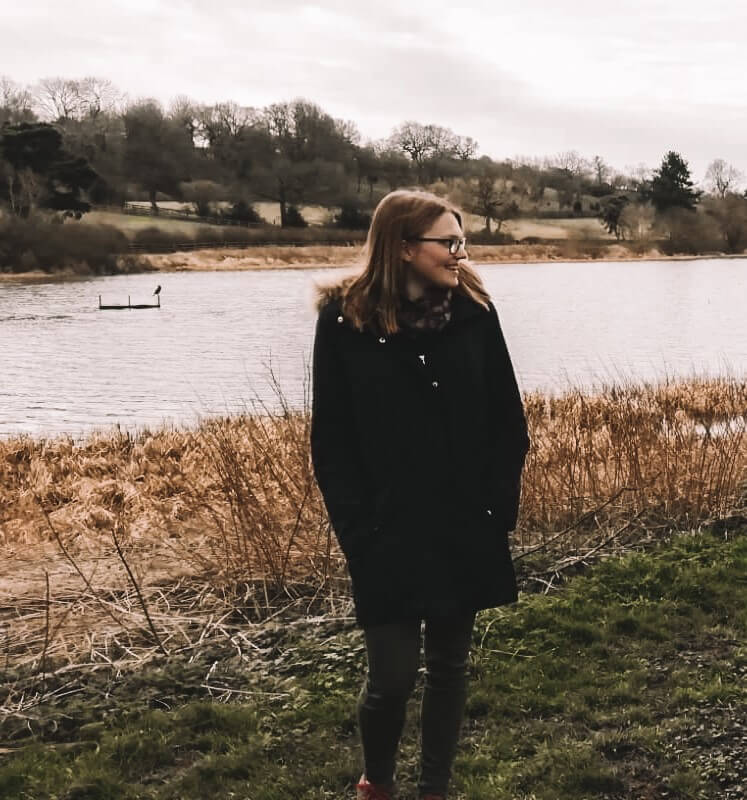 Megan Elizabeth, with blonde hair, and glasses in a navy blue winter coat is standing on a field in front of a lake