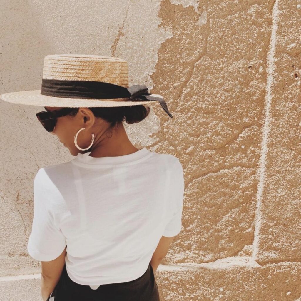 Studio Kynd founder Niobe has her back turned to camera. Her dark hair is tied in a bun and she wears a straw sunhat with dark ribbon. Niobe wears gold hoop earring and a white t-shirt. She has sunglasses on and is in front of a light brown wall.