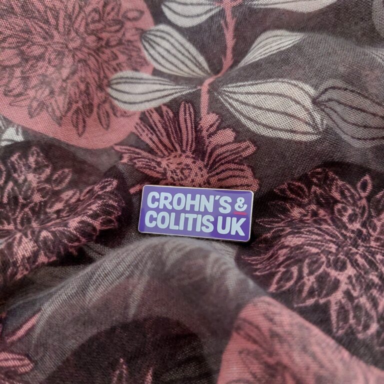 Purple Crohn's and Colitis UK pin badge on a floral fabric background to celebrate Crohn's and Colitis Awareness Week 2021