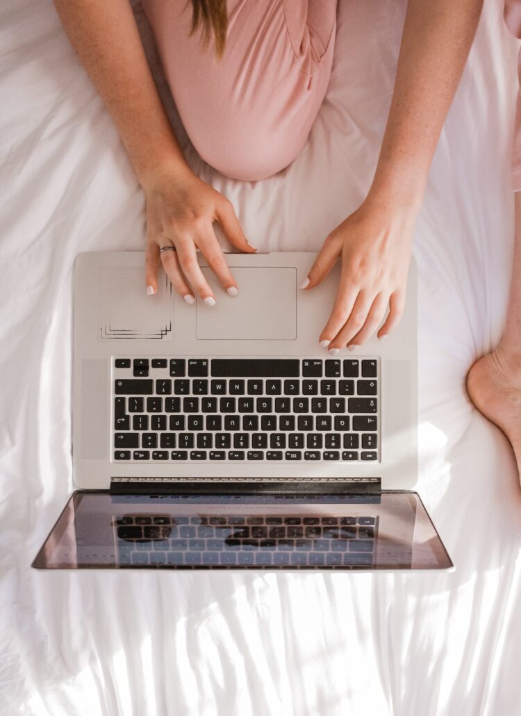 Aerial view photo of a woman in bed on her laptop. You can see her feet, hands and pink trousers.