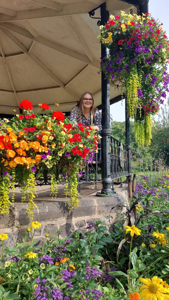 Photo of Megan, a white woman with fair hair and glasses smiling and stood on a bandstand which is covering in bright red and orange flowers.
